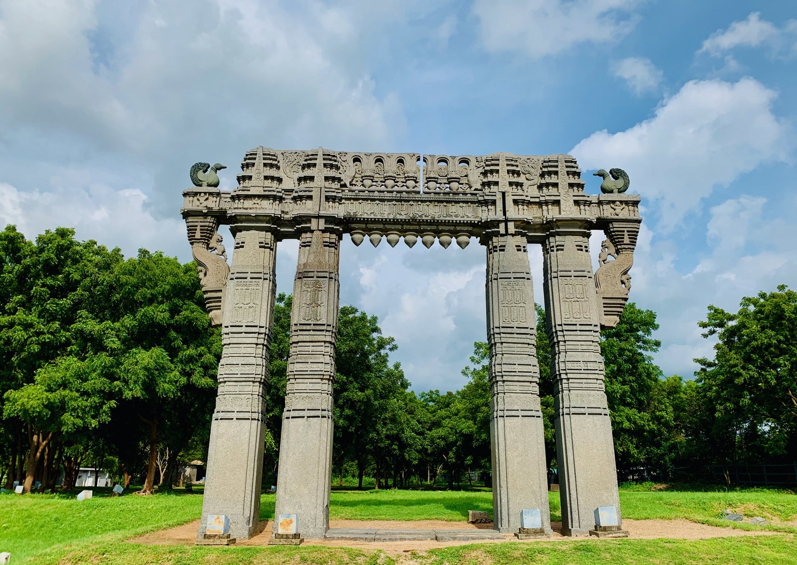 A tall stone gateway on a lawn under a blue sky. Two pairs of square carved pillars carry a heavy carved beam, with a stone swan at each upper corner.