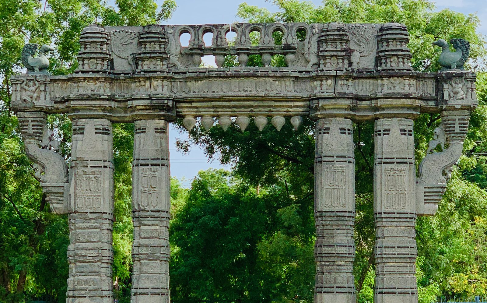The upper part of a stone gateway against trees. A row of small arches and towers runs along the top beam, stone buds hang below it, and a swan sits at each end.