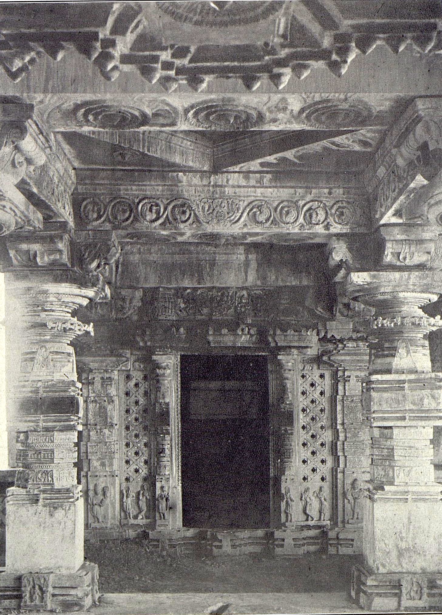 Interior of a stone temple. A carved ceiling and heavy beams frame a dark shrine doorway. Pierced stone screens stand on each side of the doorway.