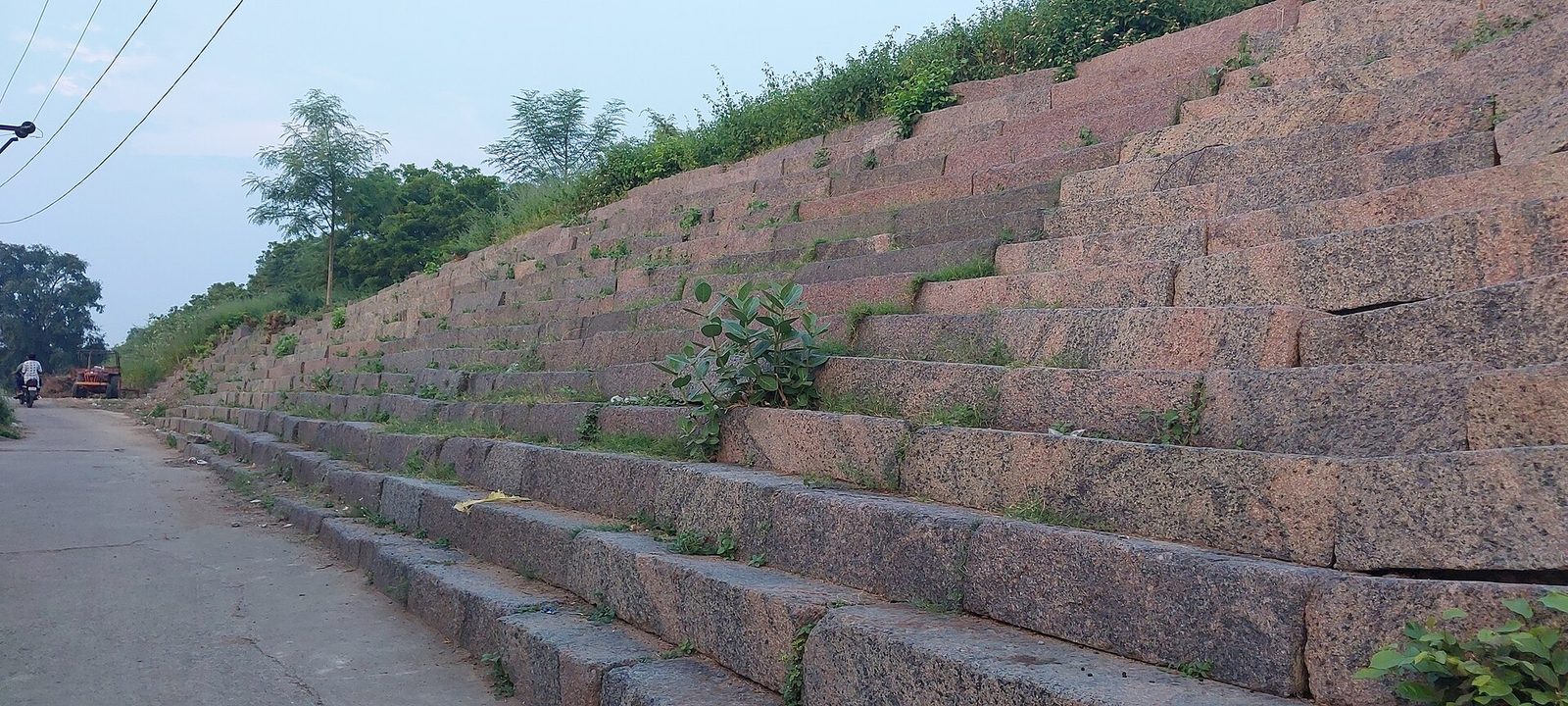A long sloping stretch of the Warangal Fort wall built of large reddish dressed stone blocks set in stepped layers, beside a road