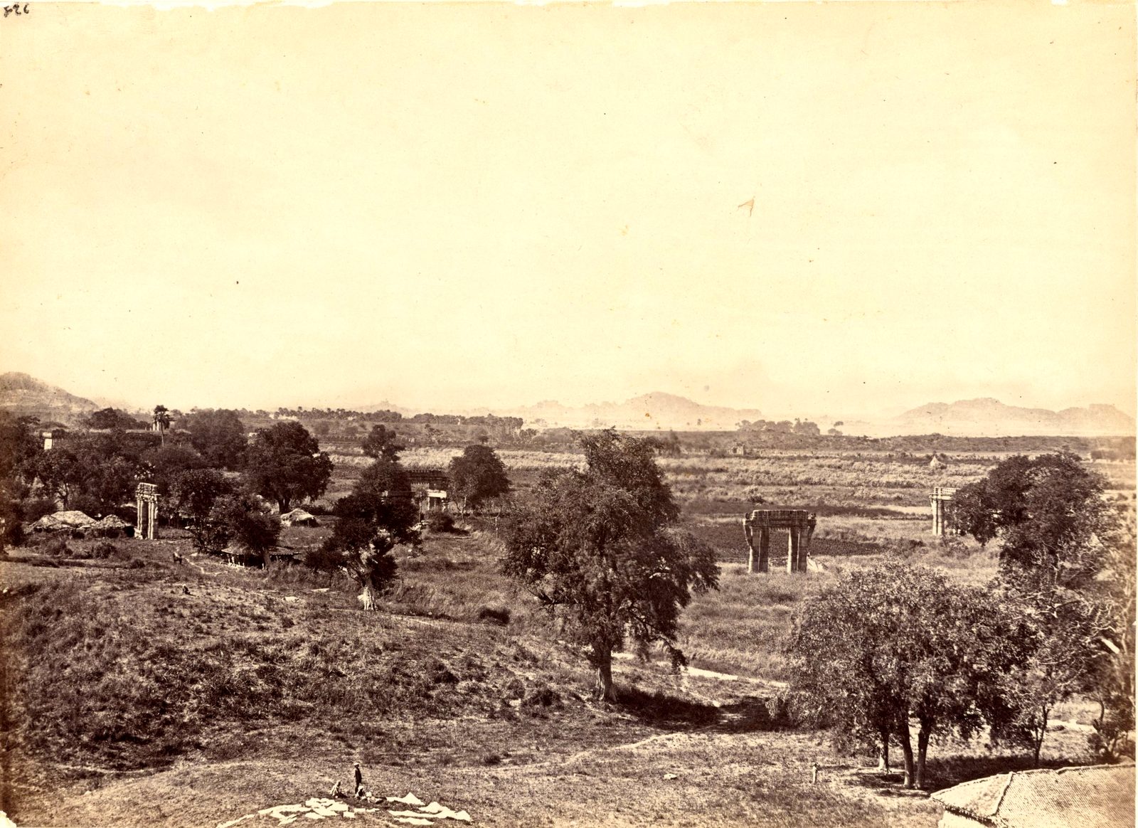 A wide view over open fields and scattered trees from a rocky hill. Several stone gateways and pillars stand alone among the fields. Low hills line the horizon.