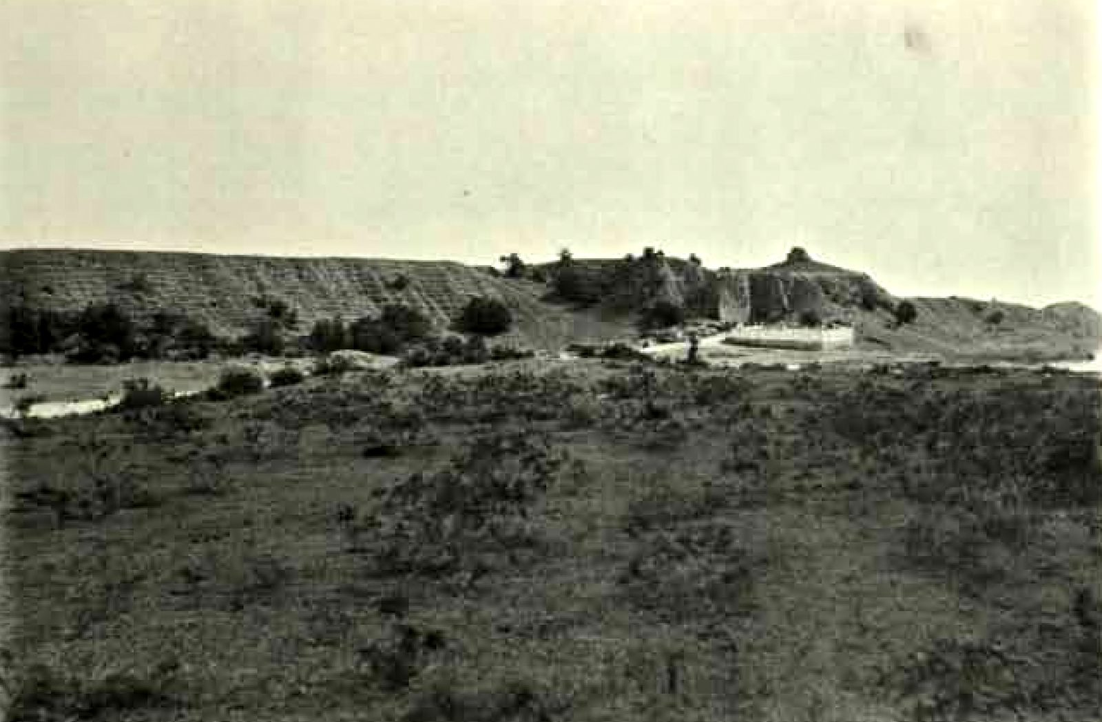 A long, low earth bank runs across the middle distance. Scrub and grass cover the open ground in front of it.