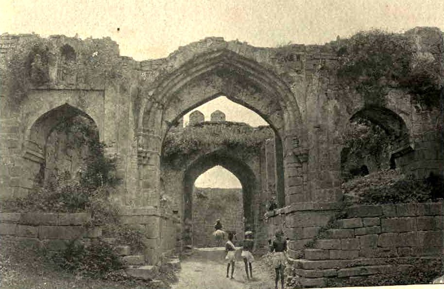 A ruined stone gateway with a tall pointed arch and a second arch behind it. Plants grow on top of the walls. Several people stand in the passage.