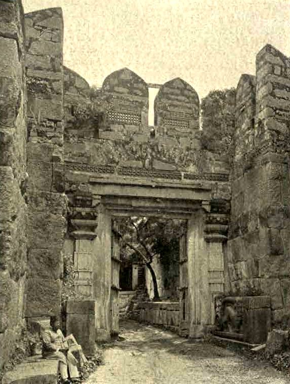 A narrow passage between high stone walls leads to a stone doorway with a flat lintel. Pointed parapets stand on top of the wall. A man sits on a stone near the doorway.