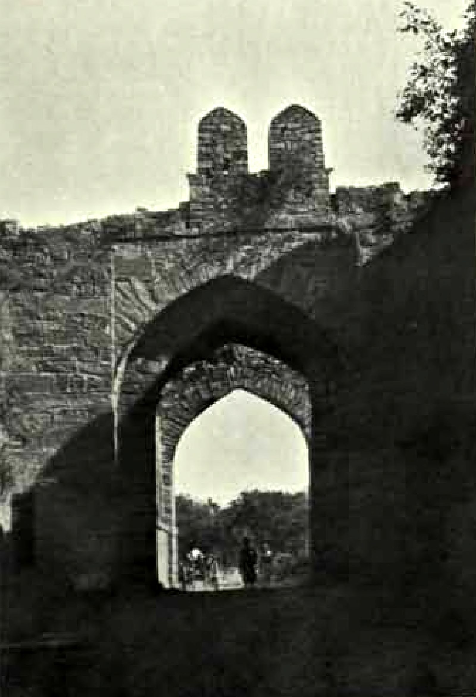 A gateway with two pointed arches in a high wall of rough stone. Two pointed stone parapets stand on top of the wall. People and a cart pass through the inner arch.