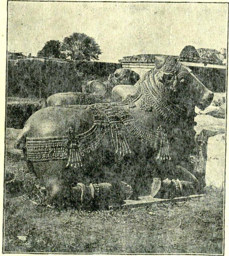 A large carved stone bull lies in grass. Rows of carved bells and chains hang around its neck. Another bull lies behind it.