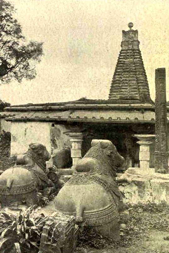 A small temple with a pale wall and a tall stepped tower on its roof. Two large carved stone bulls lie in the foreground. A tall stone pillar stands at the right.