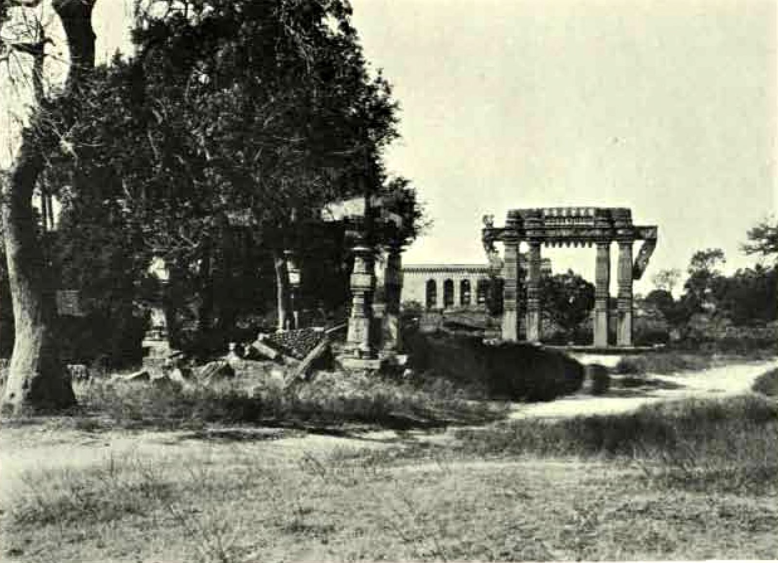 Broken carved pillars stand among large trees at the left. A carved stone gateway stands in open grassland at the right. A long building with arched openings stands behind the gateway.