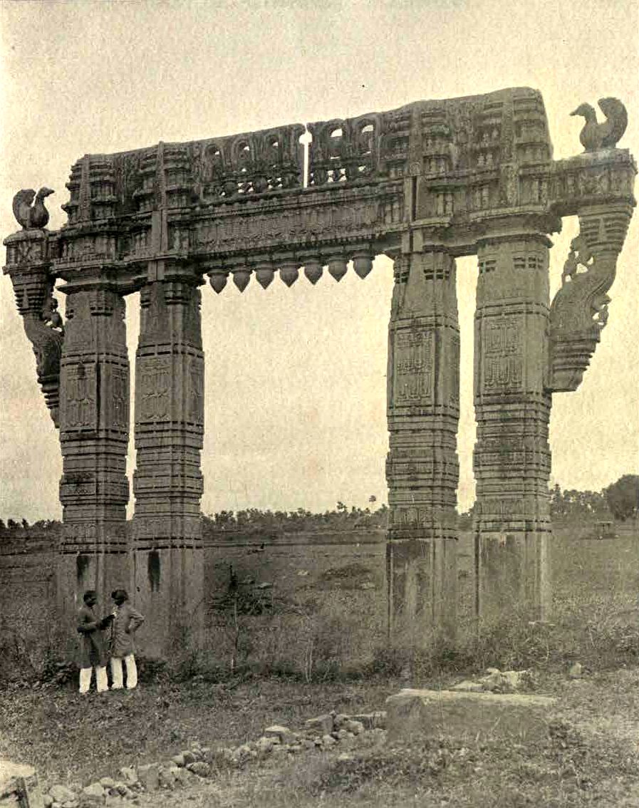 A tall stone gateway of four carved pillars and a carved crossbeam stands alone in open fields. Stone swans sit on the ends of the crossbeam. Two men stand at the foot of the left pillars.