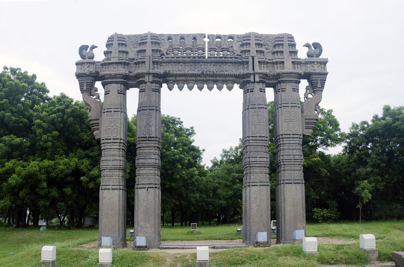 A freestanding stone gateway at Warangal Fort: two pairs of tall square pillars carrying a heavy carved lintel, with carved birds at the upper corners and curving brackets at the sides